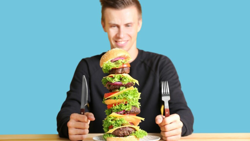 Man holding a knife and fork sitting in front of burger stacked with multiple patties and smiling.